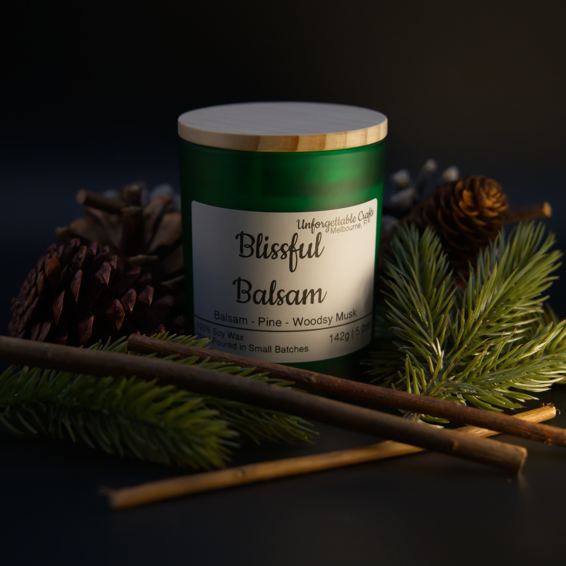 Candle labeled 'Blissful Balsam' surrounded by pine branches and cones on a dark background