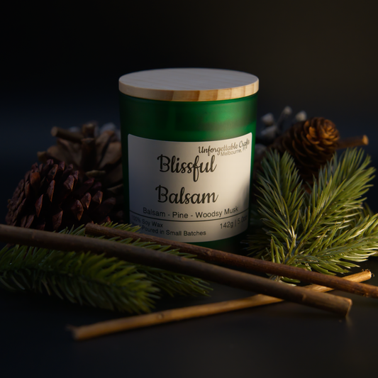 Candle labeled 'Blissful Balsam' surrounded by pine branches and cones on a dark background