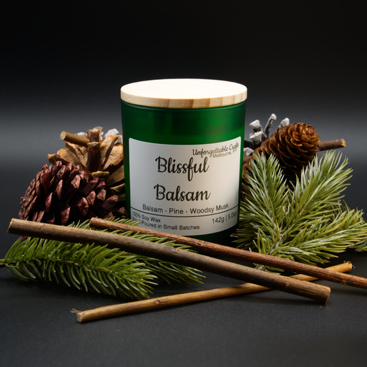 Candle labeled 'Blissful Balsam' surrounded by pine branches and cones on a dark background