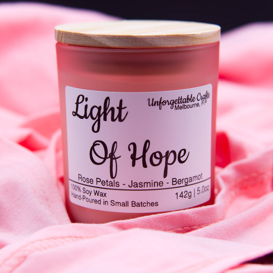 Candle labeled 'Light Of Hope' with a wooden lid on a pink fabric background