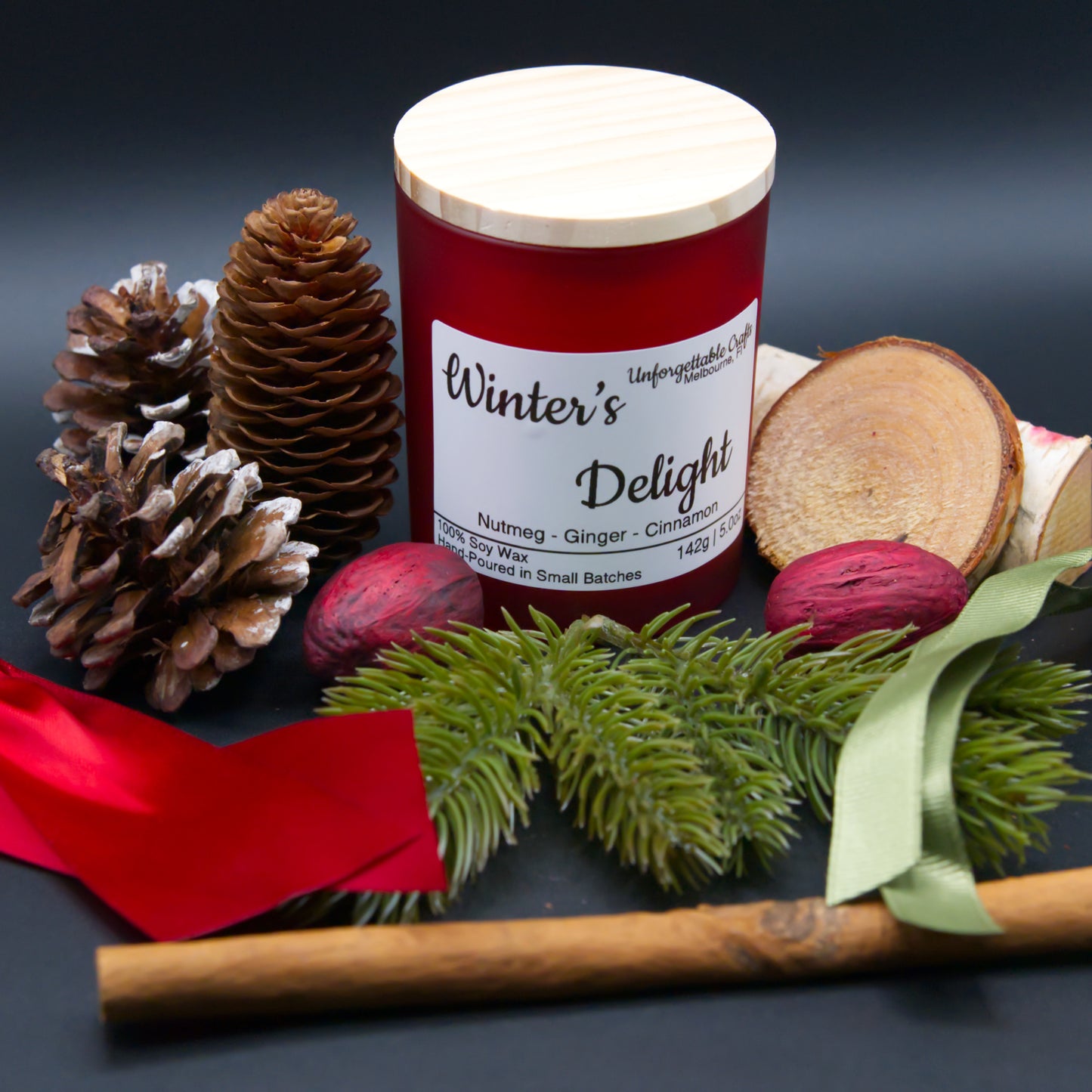 Candle labeled 'Winter's Delight' surrounded by pine cones, a cinnamon stick, and greenery on a dark background.