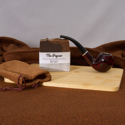Bar of soap labeled 'The Papaw' with a smoking pipe on a wooden board against a brown fabric background.